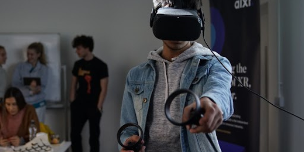 A young male wearing virtual reality googles and holding a virtual reality tool in each hand. People in the background on the left of the image